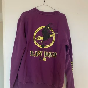 Lila crewneck Random Bastards - Säljer en lila crewneck från Random Bastards med tryck i gult och svart på ryggen och ärmen. Tröjan har rund hals, långa ärmar och är gjord i mjuk bomull. 