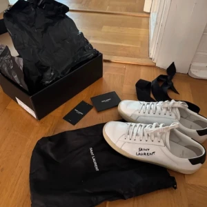 Saint Laurent vita sneakers strl 39 - Snygga vita sneakers från Saint Laurent med svart häl och handskriven logga på sidan. Klassisk låg modell med snörning och platt sula. Tillverkade i skinn och levereras med originalkartong och dustbags. Perfekta för en clean och stilren look. Pris kan diskuteras vid snabb affär.