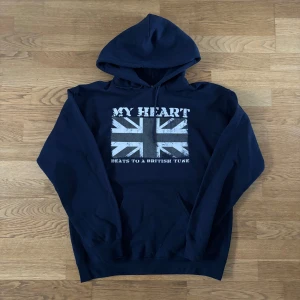 Mörkblå hoodie från Drakenberg - Snygg mörkblå hoodie med huva och känguruficka. På bröstet finns ett stort tryck med brittiska flaggan och texten 'MY HEART BEATS TO A BRITISH TUNE'. Perfekt för dig som gillar streetstyle och vill sticka ut lite extra.