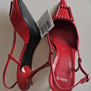 Röda pumps med spetsig tå från MNG - Helt nya, Snygga röda pumps från MNG med lackad finish, spetsig tå och dekorativt spänne med nitar framtill. Skorna har smala remmar och hög klack som ger en elegant siluett. Priset kan sänkas vid snabbaffär. Storlek 39 på de och köptes nypris för 599kr.