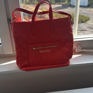 Röd quiltad axelväska från Valentino - Säljer en snygg röd axelväska från Valentino med quiltad design och guldfärgade dragkedjor. Väskan har både handtag och axelrem, samt en praktisk ytterficka med dragkedja och flera fack inuti. Perfekt för dig som vill ha plats för allt och ändå se trendig ut.