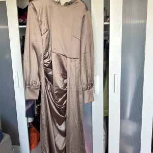 Champagnefärgad långklänning i satin - Supersnygg långklänning från shien  i champagnefärgad satin. Klänningen har långa ärmar med breda manschetter, draperad detalj i sidan och en elegant, rak siluett. Perfekt för dig som vill sticka ut med en stilren och lyxig look.