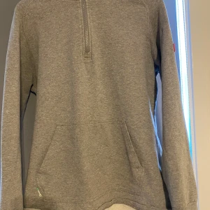 Grå hoodie från Huski Wear med half zip - Grå hoodie från Huski Wear med broderad logga på huvan och röd patch på ärmen. Tröjan har half zip framtill, känguruficka och är tillverkad i mjuk bomullsblandning. Perfekt för chill dagar eller när du vill ha en clean och enkel stil.