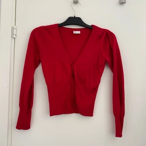 Röd kofta med v-ringning och knappar - Snygg röd kofta med v-ringad hals och knäppning framtill. Modellen är figurnära med långa ärmar och ribbade muddar. Perfekt att slänga över en topp för en färgklick i din outfit.