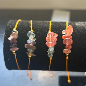 Färgglada armband med stenar - Fyra armband med orange snöre och oregelbundna pärlor i rosa, blått och lila. Varje armband har justerbar knytning och små guldfärgade detaljer. Stenarna ger en unik och bohemisk vibe, perfekt för att addera färg till din stil.