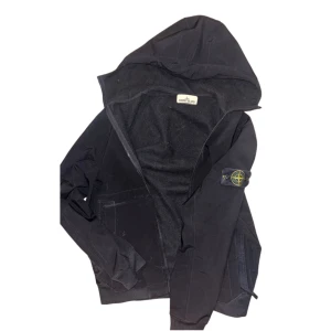 Svart softshelljacka från Stone Island - Svart softshelljacka från Stone Island med huva och ikonisk logga på ärmen. Jackan är tillverkad i stretchig polyester med vattenavvisande membran och mjuk fleece på insidan. Stängs med dragkedja och har praktiska fickor framtill. Perfekt för dig som gillar stilren och funktionell streetwear.