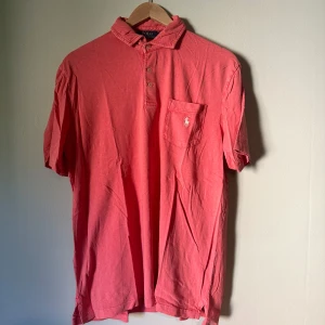Rosa pikétröja från Polo Ralph Lauren - Snygg rosa pikétröja från Polo Ralph Lauren med klassisk krage, tre knappar och bröstficka med broderad logga. Tillverkad i mjuk bomull och har korta ärmar. Perfekt för en avslappnad men stilren look. Storlek M
