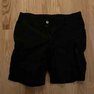 Svarta shorts från Sail Racing - Svarta shorts från Sail Racing med klassisk passform och flera fickor, inklusive bakficka med logga. perfekta för sommardagar, men även till arbetsmiljöer. Stängs med knapp och dragkedja. Snygga och stilrena shorts för en avslappnad look. Lite liten i storlek, men en XS