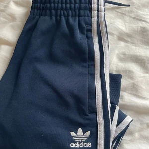 Mörkblå Adidas mjukisbyxor  - Mörkblå mjukisbyxor från Adidas med klassiska vita ränder längs sidorna och broderad logga framtill. Byxorna har resår i midjan och muddar vid bensluten💙