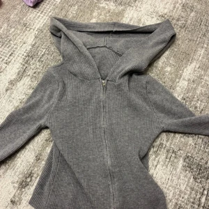 Grå ribbad hoodie med dragkedja - Säljer en grå ribbad hoodie med dragkedja framtill och huva. Tröjan har lång ärm och är tillverkad i ett mjukt, stickat material som känns både chill och stilren. Perfekt för lager-på-lager och passar till allt från jeans till mjukisbyxor.