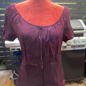 Plommonlila blus från Indiska - Supersöt plommonlila blus från Indiska med kort ärm och rundad, rynkad urringning med knytband framtill. Tunt, lätt tyg som ger en flowy känsla. Perfekt för dig som gillar romantiska och luftiga toppar.