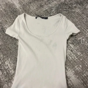 Vit ribbad topp från 157, XS/S - En stilren vit ribbad topp från 157 med korta ärmar och rundad, djup urringning. Toppen är figurnära och har en enkel, fräsch look som passar perfekt till jeans eller kjol. Materialet är mjukt och stretchigt för en bekväm känsla hela dagen.