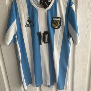 Argentina Maradona 10 Le Coq Sportif tröja - Klassisk Argentina fotbollströja med blå och vita ränder, nummer 10 och Maradona på ryggen. Broderad landslagssköld och Le Coq Sportif-logga på bröstet. Kortärmad modell i lätt och ventilerande material, perfekt för fotbollsälskare.