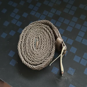 Beige flätat bälte med skinnspänne - Snyggt beige bälte med flätad design och detaljer i skinn vid spännet. Bältet har ett klassiskt metallspänne i silverfärg och passar perfekt till jeans eller chinos. Den flätade strukturen ger en cool och avslappnad vibe.