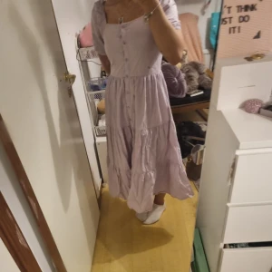 Ljuslila långklänning med knappar - Superfin ljuslila långklänning med knappar framtill och volangdetaljer. Klänningen har korta puffärmar och en söt, romantisk känsla. Är lite liten på mig.