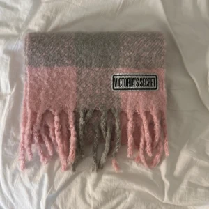 Rutig rosa och grå halsduk Victoria's Secret - Supermjuk och fluffig halsduk från Victoria's Secret i rutigt mönster med rosa och grå toner. Halsduken har långa fransar i ändarna och en tydlig logga framtill. Perfekt accessoar för att lyfta din vinterstil. Storlekarna är 228x30x0,8