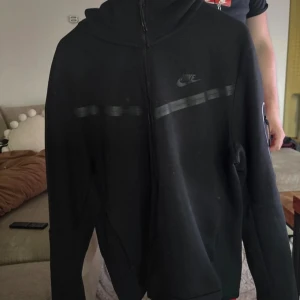 Svart Nike zip-hoodie i storlek L - Svart hoodiejacka från Nike med hel dragkedja och huva. Jackan har diskreta svarta detaljer över bröstet och Nike-logga på vänster sida. Tillverkad i mjukt bomullsmaterial, perfekt för chill eller träning. Klassisk passform med långa ärmar och fickor på sidorna.