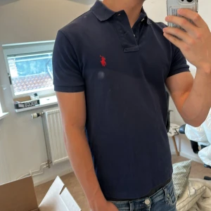 Marinblå pikétröja Polo Ralph Lauren - Säljer en marinblå pikétröja från Polo Ralph Lauren i slim fit. Tröjan har klassisk krage, korta ärmar och röd broderad logga på bröstet. Tillverkad i bomull och passar perfekt till jeans eller shorts för en clean look.