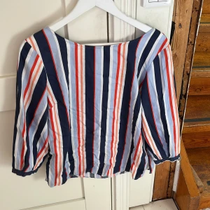 Randig blus från Hampton Republic - Snygg blus från Hampton Republic i storlek 40. Blusen har vertikala ränder i marinblått, ljusblått, vitt och rött. Den har trekvartsärmar och en liten v-ringning med knappdetalj upptill. Tillverkad i bomull, perfekt för vår och sommar.