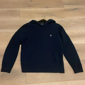 Mörkblå hoodie från Polo Ralph Lauren - Väldigt snygg mörkblå hoodie från Polo Ralph Lauren med klassisk vit broderad logga på bröstet 💯Tröjan har huva och långa ärmar otroligt bekväm, perfekt för en chill och stilren look samt mjuk insida och normal passform gör den superbekväm att bära. Köpt från kidsbrandstore och nypris ligger på 1195kr, tröjans skick är 9/10 som ny och ett galet bra pris ✅