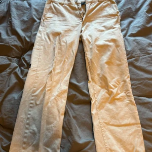 Beige cargopants från Sweet SKTBS - Snygga beige cargopants från Sweet SKTBS med raka ben och praktiska fickor på sidorna. Byxorna är tillverkade i bomull och har en klassisk cargostil med diskret logga på benet. Perfekta för en chill och streetinspirerad look.