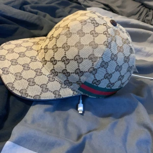 Gucci keps med GG-mönster, beige M - Exklusiv keps från Gucci i beige med klassiskt GG-monogram över hela kepsen. Grön och röd rand på sidan, justerbar rem bak i brunt skinn. Tillverkad i bomullsmix med detaljer i skinn. Perfekt för dig som vill sticka ut med lyxig streetstyle.