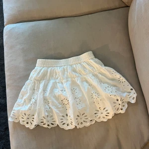 Vit kortkjol med broderade blommor - Supersöt vit kortkjol med elastisk midja och broderade blommor längs hela kjolen. Kjolen har ett par inner shorts under. Kjolen har en luftig känsla och fina utskurna detaljer i blommönster längst ner. Perfekt för en fräsch och somrig look! Strl 11-12 år. Kjolen är från Zara 