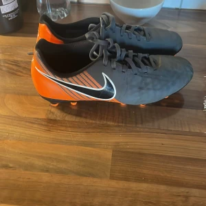 Nike fotbollsskor svart och orange - Snygga fotbollsskor från Nike i svart och orange med klassisk vit swoosh på sidan. Skorna har snörning och rund tå, samt dobbar för bra grepp på planen. Tillverkade i syntetmaterial och designade för att sticka ut på fotbollsplanen.
