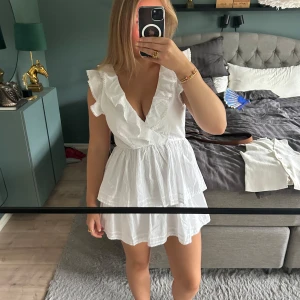 Vit volangklänning med öppen rygg - Supersöt vit kortklänning från Nelly med volanger, omlott framtill och öppen rygg med knyt. Klänningen är ärmlös och har en luftig, somrig känsla i linne. Perfekt för varma dagar när du vill känna dig extra fin.