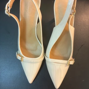 Beige pumps med spetsig tå och klack - Snygga beige pumps med spetsig tå och smal klack. Skorna har lackad finish, justerbara remmar med guldfärgade spännen och öppen häl. Perfekta för dig som gillar stilrena och eleganta skor med en modern touch. Helt nya endast testade där de var för små. 