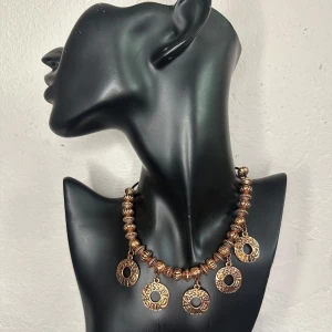 Vintagehalsband i bronsfärg med medaljonger – Retro 70-talsstil Boho Fint skick  - Rustikt statementhalsband i varm bronsfärg med präglade medaljonger och stora metallpärlor. Inköpt second hand som auktionsfynd från ett smyckeparti i 50–70-talsstil. Justerbar längd, ca 49 cm. I fint skick med jämn patina – ett lätt men kraftfullt smycke som lyfter vilken enkel outfit som helst.  Ingen prutning – men jag erbjuder gärna bundlerrabatt. Rensar smyckesskrinet – kika gärna på mina andra annonser också!  