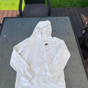 Vit hoodie från Nike med logga - Säljer en stilren vit hoodie från Nike med svart broderad logga på bröstet. Tröjan har huva, lång ärm och en stor magficka framtill. Perfekt för dig som gillar en clean och sportig look.