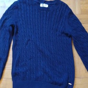 Mörkblå stickad tröja holly&White - Kabelstickad mörkblå tröja från Bold White med rund halsringning och långa ärmar. Tröjan har ett klassiskt mönster och ribbade muddar vid ärmslut och nederkant. Perfekt för lager-på-lager och chill höst- eller vinterstil.