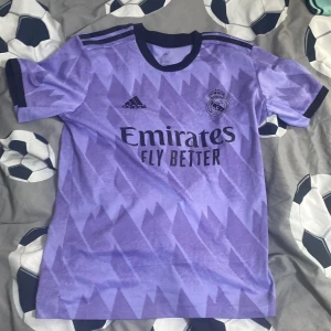 Lila Real Madrid fotbollströja Adidas - Snygg lila Real Madrid fotbollströja från Adidas med svart tryck och mönster. Tröjan har korta ärmar, svarta detaljer vid krage och ärmslut samt klubbmärke och sponsortryck på bröstet. På ryggen står det 'ADAMO' och nummer 7. Perfekt för fotbollsälskare!