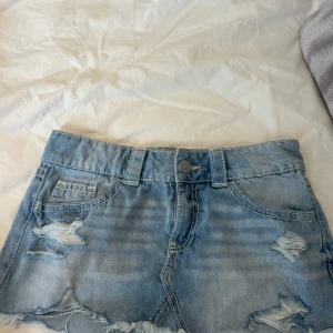Ljusblå jeanskjol med slitningar - Snygg ljusblå jeanskjol med slitna detaljer och fransig nederkant. Klassisk femficksmodell med bälteshällor och dragkedja fram. Perfekt för dig som gillar en avslappnad och trendig stil.