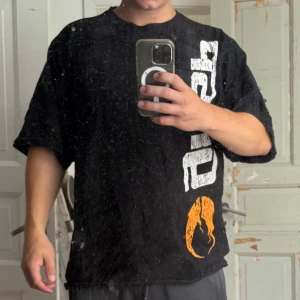 Svart oversized t-shirt med tryck - Svart oversized t-shirt med stort vitt tryck längs sidan och en orange grafisk detalj nedtill. T-shirten har en avslappnad passform och korta ärmar, perfekt för en chill och streetinspirerad look.