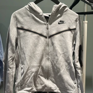 Grå hoodie från Nike med dragkedja - Snygg grå hoodie från Nike med hel dragkedja och huva. Tröjan har svarta detaljer, två fickor och Nike-logga på bröstet. Perfekt för chill eller träning, med en clean och sportig look.
