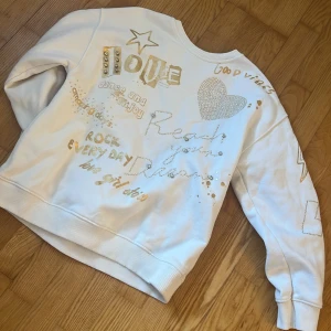 Vit sweatshirt med guldtryck från Zara - Vit sweatshirt från Zara med coola guldiga tryck, paljettdetaljer och stenar. Text och motiv som stjärnor, hjärta och peppiga budskap på framsidan. Mjuk insida och ribbade muddar. Perfekt för dig som gillar statement-plagg och snygga detaljer. Säljer i storlek 11-12 typ ungefär som xxs💞