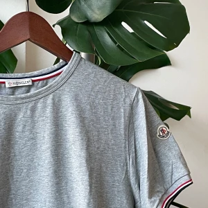 Moncler t-shirt  - Snygg grå t-shirt från Moncler i slim fit-modell. T-shirten har korta ärmar, rund halsringning och Moncler-logga på ärmen. Röd, vit och blå randig detalj på insidan av kragen och vid ärmslut. Perfekt för en clean och stilren look.