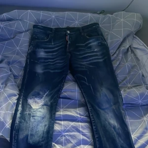 Dsquared2 blå slitna jeans herr - Säljer ett par blå jeans från Dsquared2 med coola slitningar och tvättade detaljer. Jeansen har klassisk femficksdesign, grönt Dsquared2-läderpatch bak och smal passform. Perfekta för dig som gillar streetstyle och vill sticka ut lite extra.