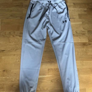 Grå mjukisbyxor från Under Armour - Snygga grå mjukisbyxor från Under Armour med svarta detaljer och dragsko i midjan. Byxorna har resår vid bensluten och praktiska fickor med dragkedja. Perfekta för chill eller träning.