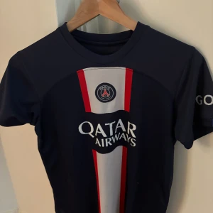 PSG Neymar Jr matchtröja blå - Paris Saint-Germain fotbollströja i mörkblått med vita och röda detaljer. Neymar Jr och nummer 10 tryckt på ryggen, Qatar Airways-logga på bröstet och 'GOAT' på ärmen. Tillverkad i lätt och ventilerande material, perfekt för fotboll eller att bära som supporter.