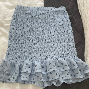 Ljusblå blommig volangkjol H&M - Supersöt ljusblå kortkjol från H&M med små svarta blommor och volangkant nertill. Kjolen är smockad och har en skön, stretchig passform. Perfekt för dig som gillar en feminin och trendig stil. Materialet är 100% polyester.