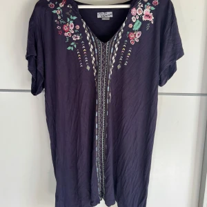 Lila blommig blus från Atlas for Women - Supersöt lila blus med v-ringning och broderade blommor i rosa, rött och grönt vid axlarna. Blusen har korta ärmar och ett dekorativt mönster längs med framsidan. Perfekt för dig som gillar färg och detaljer!