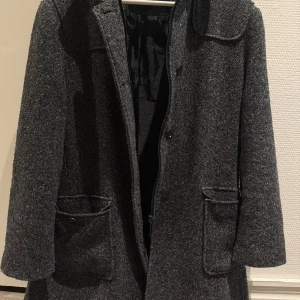 Mörkgrå ullkappa med stora fickor - Snygg mörkgrå kappa från Kappahl i ullmix med klassisk krage och stora fickor framtill. Stängs med både dragkedja och knappar. Fodrad insida för extra komfort.