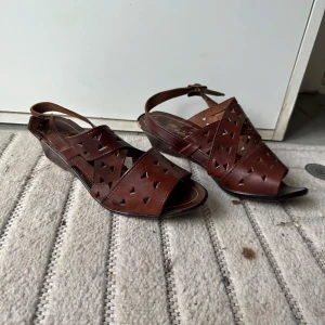 Bruna italienska sandaler i skinn - Snygga bruna sandaler i äkta skinn med öppen tå och låg klack. De har coola utskurna triangeldetaljer och justerbart spänne vid ankeln. Perfekta för varma dagar när du vill ha en klassisk och stilren look.