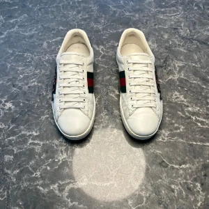 Gucci ace med ormbrodyr - Snygga vita sneakers från Gucci i skinn med klassiska gröna och röda ränder på sidorna och broderad orm i rött och svart. Baksidan har metallicdetaljer i grönt och rött med Gucci-logga. Platt sula och snörning framtill. Perfekta för dig som vill sticka ut. Väldigt bra skick på skorna 👌.         Priset kan självklart diskuteras!