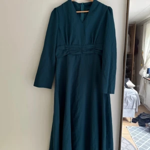 Mörkblå långklänning med v-ringning - Snygg mörkblå långklänning med v-ringad hals och långa ärmar. Klänningen har markerad midja med veckade detaljer och dragkedja bak. 