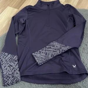 Lila tränings-tröja SOC half zip - Sportig lila långärmad tränings-tröja från SOC med half zip och coolt vitt mönster på ärmsluten. Tillverkad i mjukt och stretchigt syntetmaterial som andas. Perfekt för dig som gillar att röra på dig och vill ha en snygg och funktionell look.