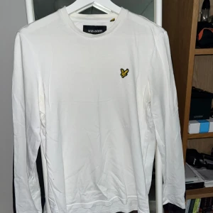Vit långärmad tröja från Lyle & Scott - Vit långärmad tröja från Lyle & Scott med klassisk rund hals och ikonisk gul fågel-logga broderad på bröstet. Tillverkad i mjuk bomull som känns skön mot huden. Perfekt för en clean och stilren look.
